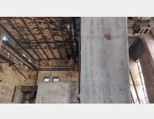 Innenraum einer verlassenen Industriehalle mit beschädigter Decke aus Stahlträgern und bröckelndem Putz. Backsteinwände, Betonpfeiler und alte Rohre bilden einen heruntergekommenen Fabrik-Look, während Licht durch kleine Fenster eindringt.