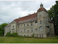 Verfallenes Herrenhaus mit grauer Fassade, roten Ziegeldächern und Turmaufsatz. Das Gebäude steht inmitten einer Wiese, umgeben von Bäumen, mit erkennbaren Schäden wie bröckelndem Putz und beschädigten Fenstern.