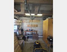 Innenansicht einer kleinen Werkstatt oder Abstellkammer mit freiliegenden Holzbalken und Deckenleuchten, einer Werkbank mit Werkzeugleisten an einer rau verputzten Wand sowie aufgehängten Kabelrollen. Links stehen Regale mit Kartons, rechts ein hoher Holzschrank und ein runder Grill; am Boden liegen Geräte und diverse Gegenstände.