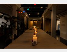 Langer Holztisch in einem dimm beleuchteten Raum, dekoriert mit brennenden Kerzen, Tannenzapfen und roten Kugeln; bunte Lichterketten hängen an der Decke. Im Hintergrund stehen Pflanzen und vereinzelte Personen.