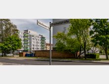 Straßenecke mit den Straßenschildern „Hückelhovener Ring“ und „Freiheitsstraße“ im Vordergrund, umgeben von Bäumen. Im Hintergrund mehrstöckige Wohngebäude mit Balkonen sowie ein gepflasterter Gehweg und eine niedrige Mauer.