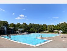 Ein großes Freibad mit mehreren rechteckigen Schwimmbecken, umgeben von einer gepflasterten Fläche. Im Hintergrund sind grüne Bäume, eine Rutsche und ein Turm zu sehen, der Zugang zu den Rutschen bietet.