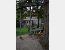 Großer Garten mit einem schattigen Sitzbereich unter einem Baum, ausgestattet mit einem Holztisch und Stühlen, neben einer Schaukel aus Seilen. Im Hintergrund steht ein zweigeschossiges Haus mit hellem Putz, rotem Dach und großzügigem Gartenbereich.