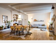 Helles Wohnzimmer mit offener Gestaltung, einem großen Holztisch mit gepolsterten Stühlen im Vordergrund und einer grauen Couch im Hintergrund. Der Raum hat helle Wände, sichtbare Deckenträger, Holzböden, Pflanzen und minimalistische Dekoration wie schwebende Regale und moderne Beleuchtung.