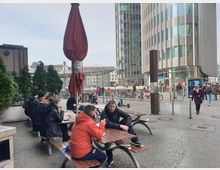 Eine städtische Platzszene mit modernen Gebäuden, darunter ein hohes, abgerundetes Bürogebäude mit Glas- und Metallfassade. Im Vordergrund befinden sich Sitzbänke mit Menschen, die essen und sich unterhalten, sowie ein geschlossenes rotes Sonnenschirm. Fahrräder stehen in Reihung, und Fußgänger bewegen sich durch den Platz.