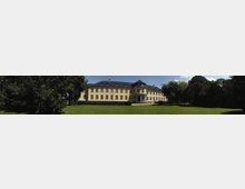 Panoramabild eines großen, gelb gestrichenen Gebäudes mit Walmdach und zahlreichen Fenstern, im Zentrum mit einem halbrunden Eingang mit Säulen. Im Vordergrund eine gepflegte Rasenfläche, umgeben von hohen Bäumen.