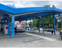 Bushaltestelle mit modernem blauen Dach und freistehenden Säulen, umgeben von einer gepflasterten Fläche. Im Hintergrund sind mehrstöckige Gebäude in Pastellfarben, Bäume und parkende Autos zu sehen.