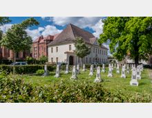 Ein kleiner Friedhof mit weißen Grabsteinen, die rote Sterne und Verzierung tragen, umgeben von Grünflächen und Bäumen. Im Hintergrund befinden sich ein historisches Gebäude mit rotem Backstein und ein einfaches, weißes Gebäude mit Satteldach.