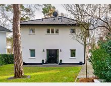 Weiße, zweistöckige Wohnhausfassade mit einem schwarzen Eingangstürrahmen, flankiert von zwei kleinen Pflanzen in Töpfen. Im Vordergrund ein Garten mit grünem Rasen, Bäumen und Büschen, und rechts ein gepflasterter Weg.