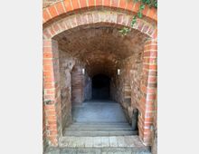 Backstein-Gewölbetunnel mit Sicht auf eine absteigende Treppe, flankiert von gemauerten Wänden mit Bögen. Oberhalb des Eingangs wächst Efeu.