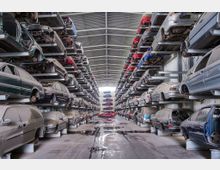 Eine große Lagerhalle mit mehreren gestapelten Ebenen von gebrauchten und beschädigten Autos, die in Metallregalen geparkt sind. Der Boden ist aus Beton und weist Wasserpfützen auf, und die Halle ist durch ein Wellblechdach und Wände abgeschlossen.