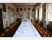 Ein eleganter Speiseraum mit einer langen, weiß gedeckten Tafel, umgeben von Holzstühlen. Die rosa gestrichenen Wände sind mit Porträts und historischen Fotografien dekoriert, während hohe Fenster, teils mit weißen Klappläden, viel Licht hereinlassen.