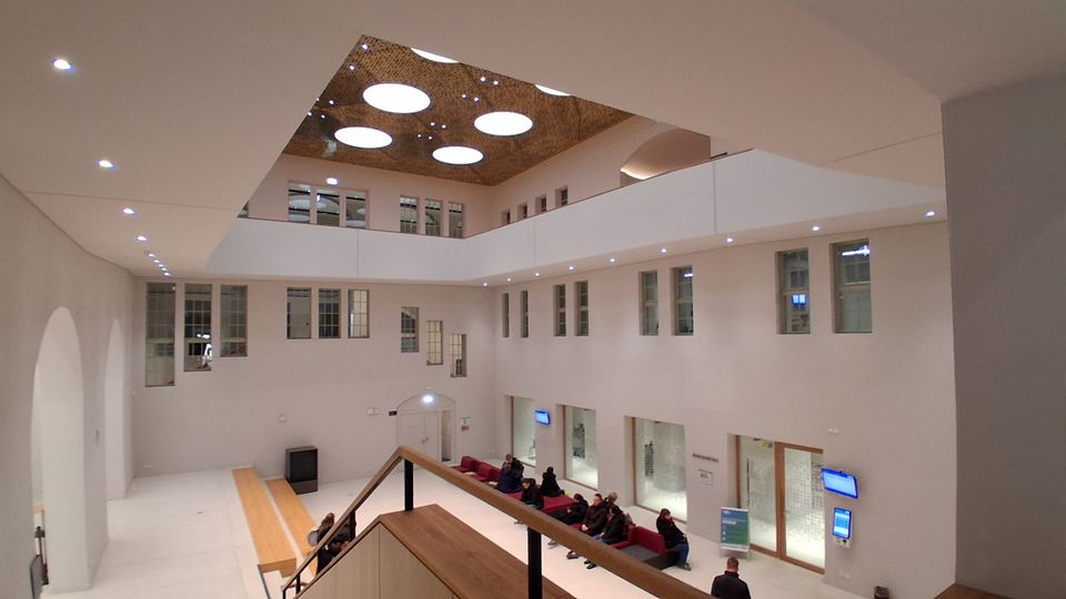 Großer, moderner Innenhof mit weißen Wänden und umlaufender Galerieebene, umgeben von zahlreichen Fenstern und Glasfronten. In der Decke befinden sich mehrere runde Oberlichter; unten ist ein Wartebereich mit Sitzbänken und digitalen Anzeigen.