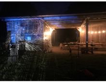 Nachtszene eines Holzunterstands, der mit warmem Licht beleuchtet ist. Auf der linken Seite wird eine Holzwand durch abstrakte, blaue Lichtprojektionen hervorgehoben, während im Unterstand Bänke und Tische erkennbar sind.