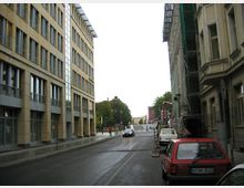 Straßenansicht in einer städtischen Umgebung mit modernen Gebäuden auf der linken Seite und älteren Gebäuden, teils mit Baugerüsten, auf der rechten Seite. Geparkte Autos stehen entlang der Straße, die in der Ferne von Bäumen und einem Denkmal gesäumt ist.