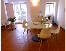 Heller Besprechungsraum mit hellem Fliesenboden, einem runden Glastisch und vier weißen Stühlen. Im Raum befinden sich ein Flipchart, ein Laptop auf einem Schreibtisch, zwei Fenster mit Tageslicht und eine Glastür mit Blick nach draußen.