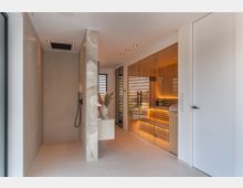 Moderner Wellnessbereich mit großformatigen Fliesen, bodengleicher Dusche mit schwarzer Armatur und einer verglasten Sauna aus Holz mit indirekter Beleuchtung. Schmale Fenster mit Lamellen, Einbauspots in der Decke und eine freistehende Trennwand aus Stein strukturieren den Raum.