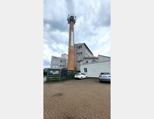 Backsteinschornstein mit Beschriftung „Luckenwalde“ neben einem grauen Gebäude im Hintergrund, umgeben von einem gepflasterten Parkplatz mit geparkten Autos und einem Metallzaun im Vordergrund, unter bewölktem Himmel.