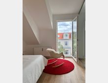 Minimalistisch eingerichtetes Schlafzimmer mit Holzfußboden, weißer Bettwäsche, einem roten runden Teppich und einem Schaukelstuhl mit Fellauflage. Ein offenes Fenster bietet Blick auf benachbarte Gebäude mit roten Dächern und Gauben.