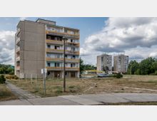 Wohnsiedlung mit Plattenbauten: Im Vordergrund ein vierstöckiges Wohngebäude mit gelben und hellblauen Balkonen, umgeben von einer unbefestigten Fläche mit vereinzelten Gehwegen. Im Hintergrund stehen höhere Plattenbauten sowie Bäume und ein Parkplatz mit geparkten Autos.