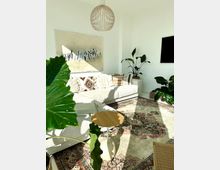 Wohnzimmer mit einem hellen, modernen Design. Ein großes, weißes Sofa mit gemusterten Kissen steht an einer Wand mit abstraktem Gemälde, umgeben von mehreren grünen Zimmerpflanzen; im Vordergrund ein orientalischer Teppich, ein kleiner Holztisch und eine hängende Lampe aus Holz.