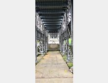 Blick durch die massive Stahlkonstruktion eines Schiffshebewerks mit sichtbaren Metallträgern und einer Betonfläche. Im Hintergrund sind eine Brücke und kleine Menschengruppen erkennbar.