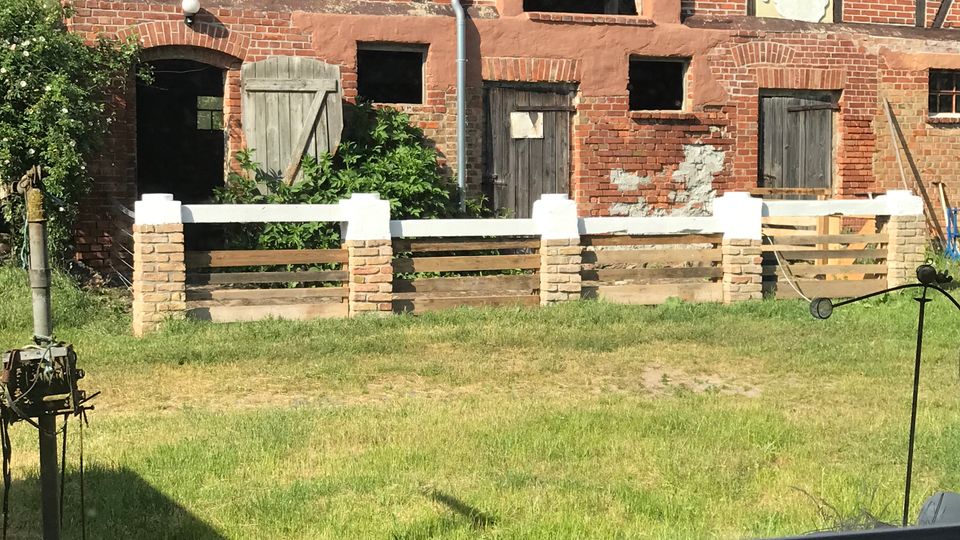 Altes Backstein-Fachwerkgebäude mit geneigtem Ziegeldach, mehreren Holz­türen und kleinen Fenstern, teils mit beschädigtem Putz. Davor liegt eine Rasenfläche mit einem niedrigen Zaun aus Steinpfeilern und Holzlatten.