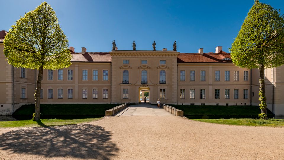 Wide view of a symmetrical historic palace facade with beige walls, red-tiled roofs, and statues along the roofline. A straight gravel path and small bridge lead to a central arched entrance, framed by trimmed trees and lawns under a clear blue sky.