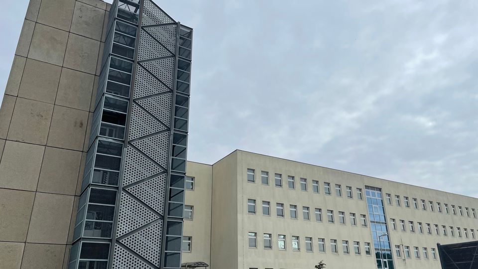 Außenansicht eines mehrstöckigen, hell verputzten Büro- oder Verwaltungsgebäudes mit vielen gleichmäßig angeordneten Fenstern und einem verglasten Mittelteil. Links ist ein angebauter, hoher Treppen- oder Aufzugsturm aus Metall und Glas mit perforierten Paneelen und diagonalen Streben zu sehen, davor ein kleiner Vorplatz mit etwas Grünfläche.