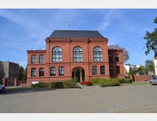 Backsteingebäude im Stil der Neorenaissance mit großen, rundbogigen Fenstern und einem Mansarddach, umgeben von einem gepflasterten Hof und Grünflächen; links und rechts sind moderne Wohnhäuser sichtbar.