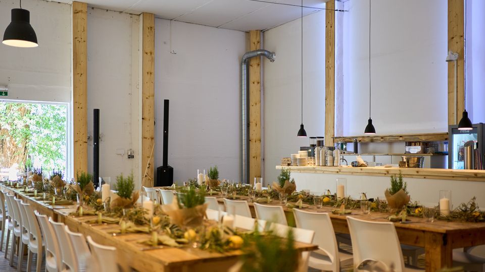Ein heller, moderner Veranstaltungsraum mit einer einfachen Holzkonstruktion, langen Holztischen und weißen Stühlen. Die Tische sind dekoriert mit grünen Pflanzen, Kerzen und Naturmaterialien, während im Hintergrund eine minimalistisch gestaltete Bar zu sehen ist.