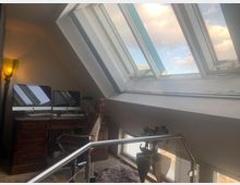 Ein Dachzimmer mit schrägen Fenstern, die helles Tageslicht hereinlassen. Im Vordergrund steht ein Holzbüroschreibtisch mit zwei Monitoren, einer braunen Bürostuhl und einer Tischleuchte, daneben ein Geländer mit Glas.