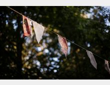 Eine Reihe von bunten Stoffwimpeln hängt an einer Schnur vor einem Hintergrund aus unscharfen, grünen Baumkronen mit Sonnenlicht, das durch die Blätter scheint.