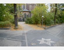 Pflasterweg mit eingelegtem Sternmuster führt zu einem Backsteingebäude mit spitzbogigen Fenstern und einem kleinen Runddach. Umgeben von Herbstbäumen und Sträuchern, liegen Laubblätter verstreut auf dem Gehweg.