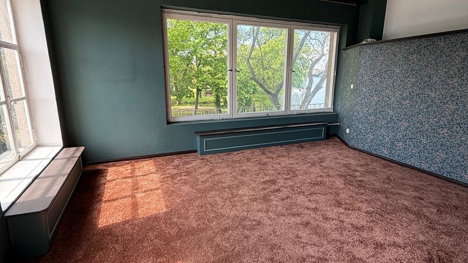 Leerer Innenraum mit dunkelgrünen Wänden, braunem Teppichboden und großen Fensterflächen, die auf Bäume im Außenbereich blicken. Links befindet sich eine Fensterbank mit eingelassenen Sitz-/Heizkörperverkleidungen, rechts eine Wand mit blau gemusterter Tapete.