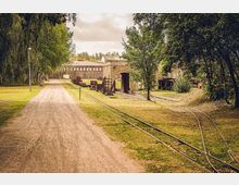 Weitläufiges Freilichtmuseum mit einem Schotterweg, alten Gleisen und historischen Backsteingebäuden. Die Umgebung ist von Bäumen gesäumt, und entlang der Gleise stehen rostige Eisenbahnwaggons und Industriemaschinen.