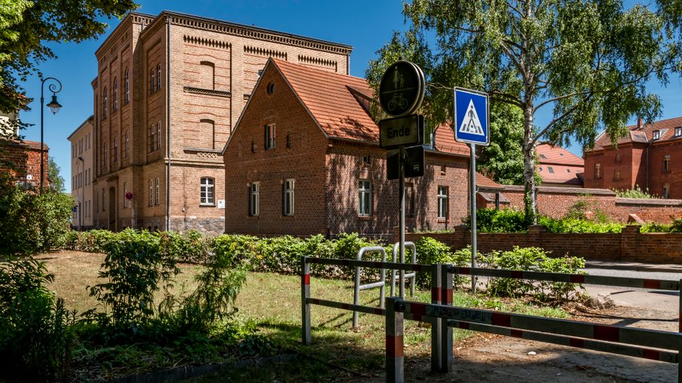 Straßenszene mit historischen Backsteingebäuden und roten Ziegeldächern, umgeben von Bäumen und niedrigen Ziegelmauern. Im Vordergrund stehen Verkehrsschilder sowie eine Absperrung an einem kleinen Weg neben einer Grünfläche.