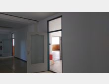 Ein Korridor mit einer geöffneten Tür zur Seite, die Milchglasscheiben enthält. Dahinter befindet sich ein heller Raum mit Fliesenboden und einer Holzvertäfelung an der Wand.