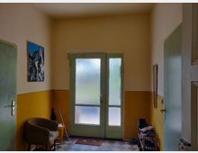 Ein kleiner Eingangsbereich mit beige-gelben Wänden und einer zweiflügeligen Glastür mit grünem Rahmen. Links steht ein Sessel, rechts ein Schuhregal mit einigen Schuhen und Zeitungen, auf dem Boden liegen Fliesen in einem Schachbrettmuster.