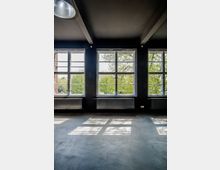 Ein leerer Innenraum mit einer dunkel gestrichenen Decke und großen Fenstern mit Metallrahmen, die einen Blick auf grüne Bäume bieten. Auf dem Boden sind Licht- und Schattenspiele sichtbar, unter den Fenstern befinden sich schmale Heizkörper.