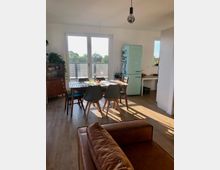 Helles Esszimmer mit Holzfußboden, einem rechteckigen Esstisch aus Holz, umgeben von vier Stühlen mit grauen und blauen Sitzflächen. Im Hintergrund befinden sich eine große Glastür zu einem Balkon, ein hellgrüner Kühlschrank und Pflanzen auf Möbeln und Fenstersimsen.