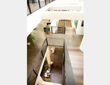 Innenansicht eines mehrstöckigen Gebäudes mit offener Galerie und Holzfußboden. Sicht auf Treppen, Geländer aus Metall und einige Pflanzen in Töpfen, mit modernen Möbeln im Erdgeschoss.