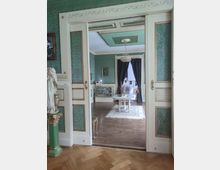 Ein historischer Innenraum mit grüner, gemusterter Tapete und weißen, verzierten Türen mit goldenen Akzenten. Im Hintergrund ist ein Raum mit einem Kronleuchter, einem weißen Tisch auf einem Teppich und dunklen Vorhängen zu sehen.