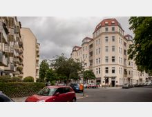 Straßenecke in einer Wohngegend mit mehrstöckigen, hellen Wohnhäusern mit roten Dächern und zahlreichen Fenstern. Im Vordergrund stehen parkende Autos, Bäume säumen die Straße, und im Erdgeschoss befindet sich ein kleines Geschäft mit Markise.