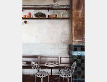 Ein gemütliches Café mit zwei weißen, filigranen Metallstühlen und kleinen runden Tischen vor einer hellen Backsteinwand. Oben an der Wand befindet sich ein rustikales Holzregal mit Pflanzen, Dekor und Kochutensilien; rechts ist ein Abschnitt mit grünen, glänzenden Kacheln sichtbar.