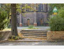 Ziegelkirche mit runden Holztüren und hohen Fenstern im Hintergrund, umgeben von herbstlichem Laub auf dem Boden. Im Vordergrund führen kleine Steinstufen, eingerahmt von niedrigen Ziegelmauern und Bäumen, zu einem gepflasterten Platz.