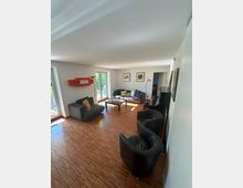 Helles Wohnzimmer mit Holzparkett, grauen Sofas und zwei schwarzen Sesseln um einen niedrigen Couchtisch. Große Fenster- und Glastüren lassen Tageslicht herein; an den Wänden hängen Bilder, daneben stehen Stehlampen und ein langes Sideboard.