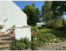 Ein Garten mit einer weißen Hauswand im Vordergrund, an der ein Holztisch mit Pflanzen steht. Rechts befindet sich eine Rasenfläche mit Bäumen und Sträuchern, im Hintergrund ist ein geparktes Auto am Rande eines Feldes sichtbar.
