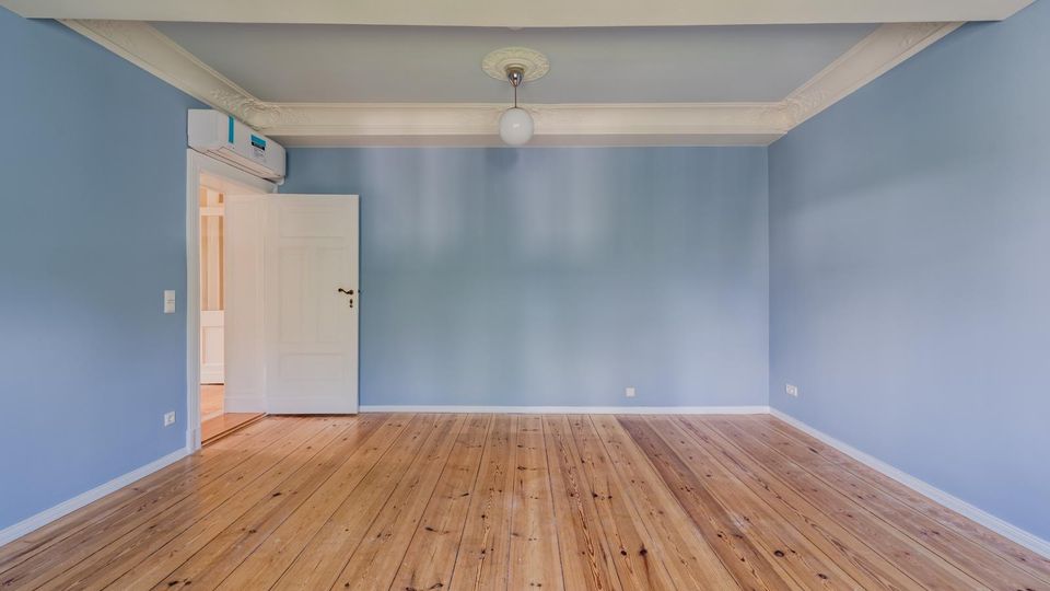 Leerer Wohnraum mit hellblauen Wänden, breiten weißen Stuckleisten an der Decke und einer zentralen Deckenlampe. Holzdielenboden und offene weiße Tür zu einem angrenzenden Flur, mit Klimagerät über der Tür.