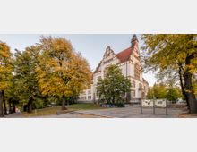 Historisches Gebäude mit rotem Satteldach und verzierten Backsteinfassaden, umgeben von Bäumen in herbstlichen Farben. Im Vordergrund befinden sich Infotafeln und ein gepflasterter Weg.